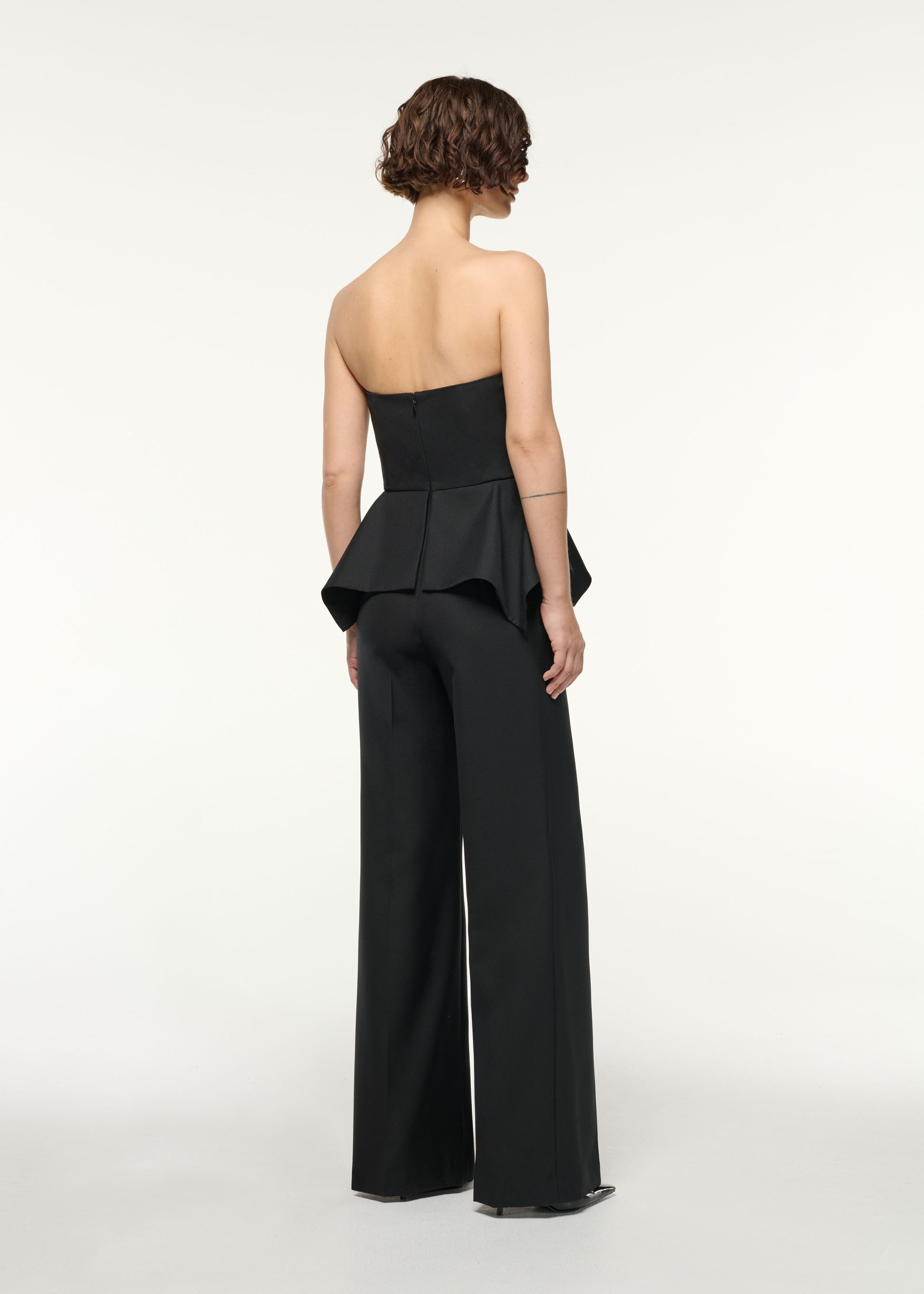 Back view of a model wearing the Crystal Button Peplum Wool Jumpsuit in Black