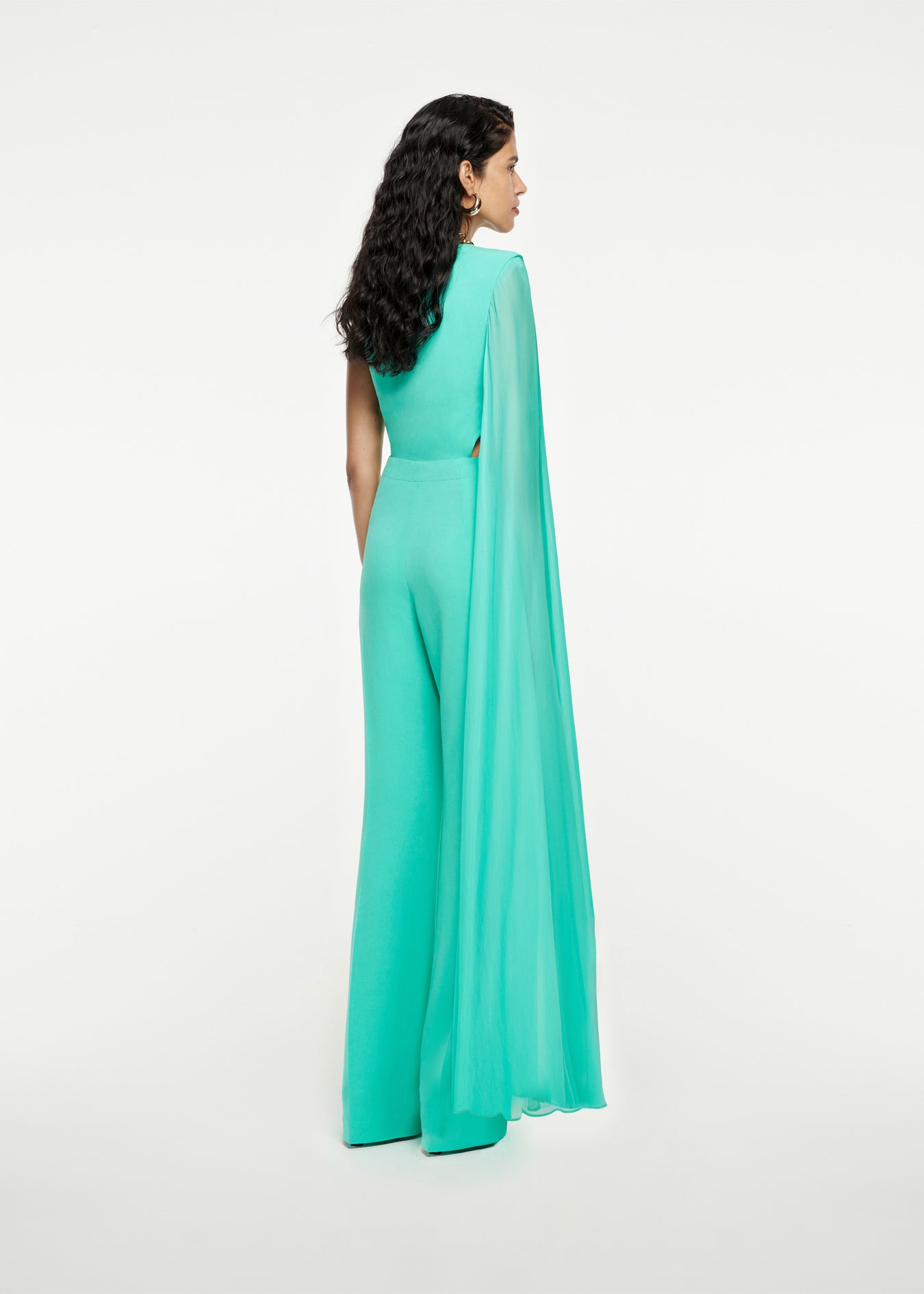 The back of a woman wearing the Asymmetric Stretch-Cady Cape Jumpsuit in Turquoise