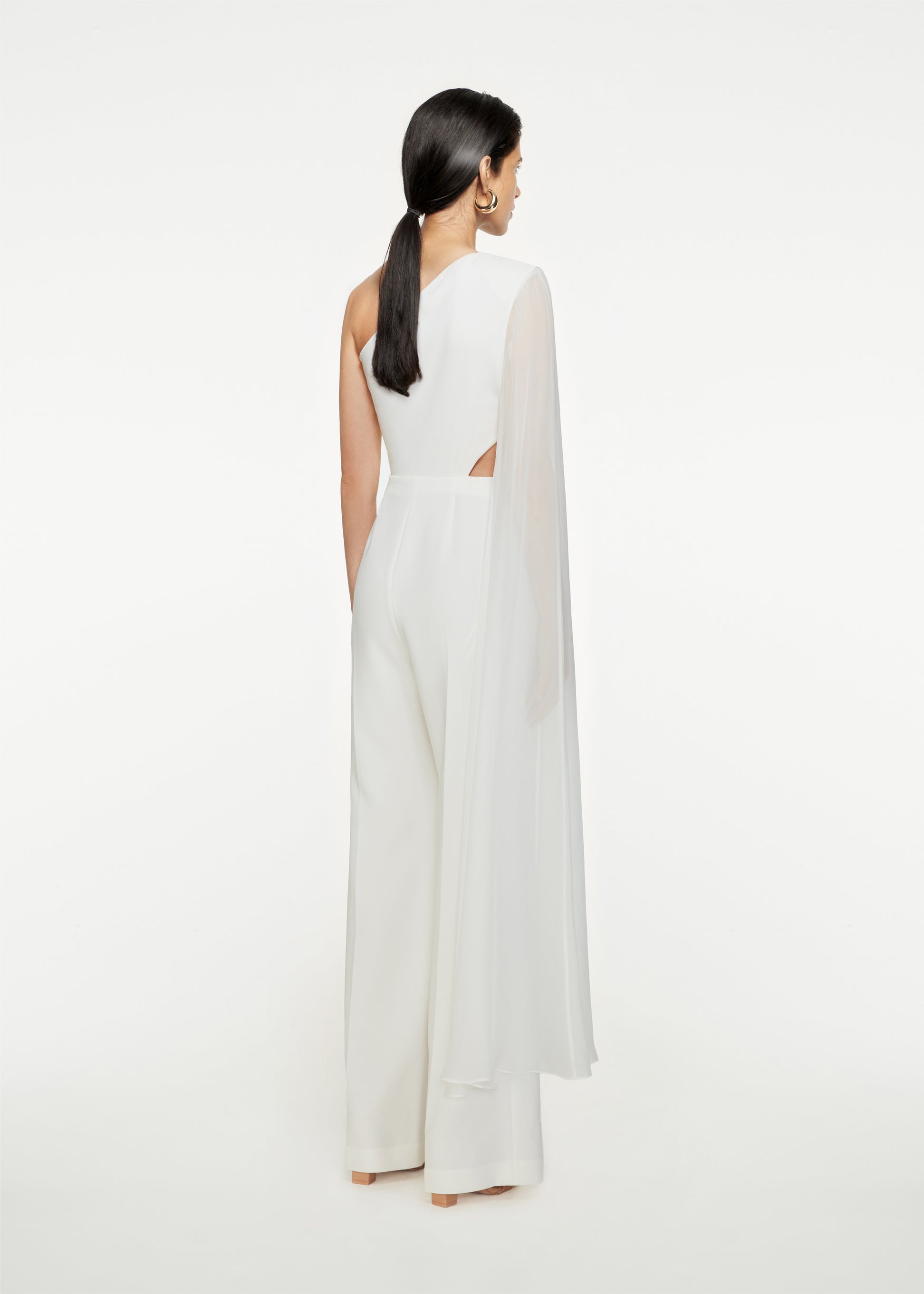 The back of a woman wearing the Asymmetric Stretch-Cady Cape Jumpsuit in White